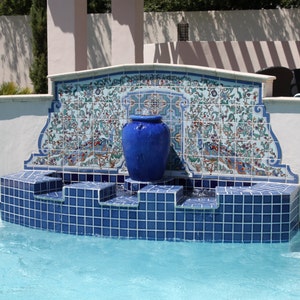 May include: A blue ceramic vase fountain with intricate tilework surrounding it. The fountain is built into a pool with blue tile and water.