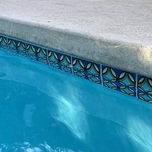 K&ouml;nnte beinhalten: Nahaufnahme des Beckenrandes eines Schwimmbades. Das Poolwasser ist ein leuchtendes T&uuml;rkisblau. Der Rand weist dekorative Fliesen mit blauen, gr&uuml;nen und schwarzen geometrischen Mustern auf, die auf einer Betonumrandung angeordnet sind.