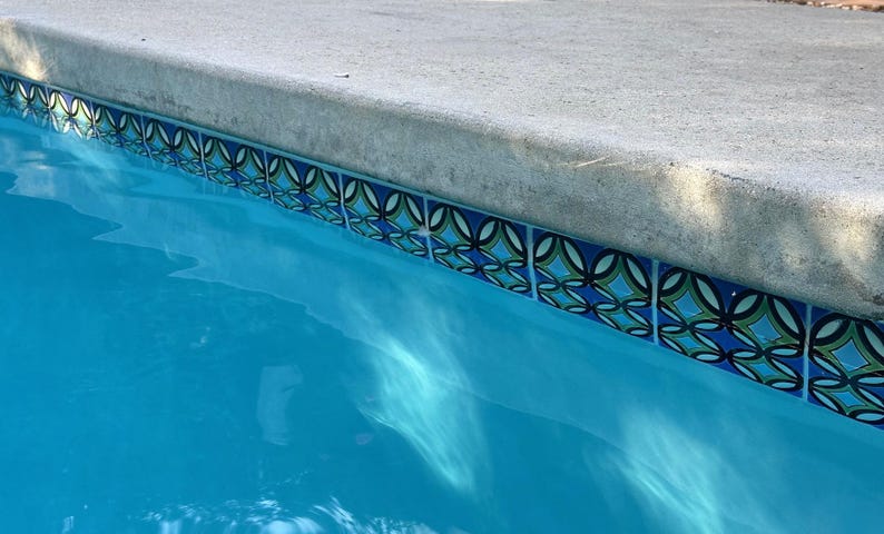 K&ouml;nnte beinhalten: Nahaufnahme des Beckenrandes eines Schwimmbades. Das Poolwasser ist ein leuchtendes T&uuml;rkisblau. Der Rand weist dekorative Fliesen mit blauen, gr&uuml;nen und schwarzen geometrischen Mustern auf, die auf einer Betonumrandung angeordnet sind.