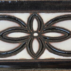 May include: Decorative ceramic tile with a glossy finish. The tile features a central white panel with a black floral design and black border. The design includes a four-petal flower and stylized scrollwork.