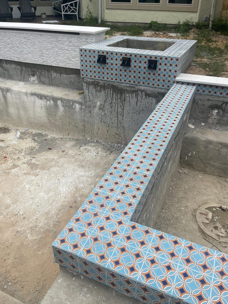K&ouml;nnte beinhalten: Eine im Bau befindliche Au&szlig;enstruktur mit einer quadratischen Feuerstelle und umliegenden Mauern. Die Oberfl&auml;chen sind mit blau, orange und wei&szlig; gemusterten Fliesen verziert. Die Struktur befindet sich in einem Betonbereich, mit einem wei&szlig;en Gesims im Hintergrund.