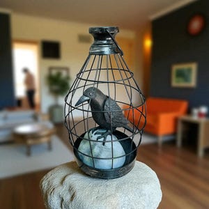 May include: A decorative black metal cage with a crow figurine inside, perched on a light blue sphere. The cage has a handle at the top and sits on a stone-like base. The crow is detailed with a textured finish.