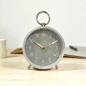 Peut inclure: Un réveil analogique gris avec une poignée en forme d'anneau doré et des pieds dorés. Le cadran de l'horloge comporte des chiffres blancs et des aiguilles dorées. L'horloge est sur une surface en bois clair.