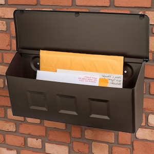 May include: A dark brown metal mailbox fixed to a red brick wall. The mailbox features a hinged lid and a rectangular body with three square decorative elements. Inside, there are envelopes and letters.