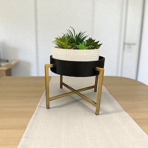Puede incluir: Un soporte decorativo para plantas con una estructura de metal negro y dorado. Sostiene una maceta blanca y negra llena de suculentas verdes. El soporte está sobre un camino de mesa de color claro.