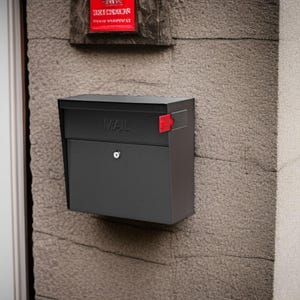 May include: A black mailbox mounted on a textured stone wall. The word "MAIL" is embossed on the front. A red flag and a small lock are visible. A red sign is above the mailbox.