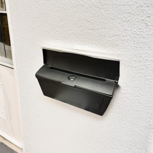 May include: A black mailbox mounted on a textured white wall. The mailbox has a hinged lid and a slot for mail. The design is modern and minimalist, suitable for residential use.
