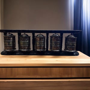 May include: Five black prayer wheels with gold and silver accents are mounted on a black wooden stand. The prayer wheels are decorated with intricate patterns and symbols.