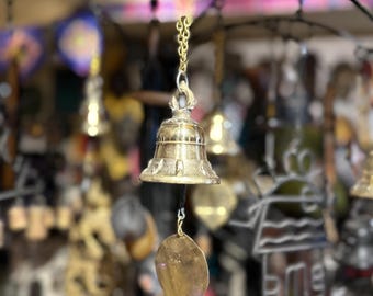 Puede incluir: Una campana dorada colgando de una cadena, con un badajo en forma de hoja. La campana tiene un diseño detallado alrededor de la base y está suspendida contra un fondo borroso de otras campanas y objetos decorativos.
