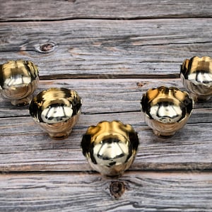 May include: Five small, gold-colored bowls with a scalloped rim are arranged on a weathered wooden surface. The bowls have a reflective, shiny interior, and the light creates highlights and shadows.