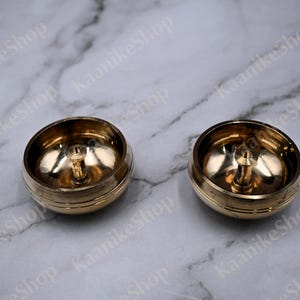 May include: Two brass-colored, bowl-shaped objects with a central, vertical post. The objects have a rounded base and a polished, reflective interior. The background is a light gray marble-like surface.