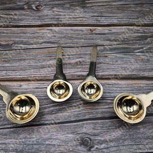 May include: Four metal ladles with a silver handle and a gold-colored bowl are arranged on a wooden surface. The ladles are designed with a rounded bowl and a curved handle, suggesting a kitchen or serving utensil.