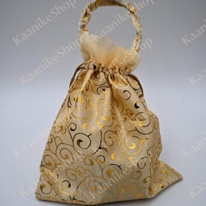 May include: A small, beige drawstring gift bag with a gold swirling pattern. The bag has a matching handle and is made of a sheer, shimmering fabric. The bag is closed with a gold drawstring.