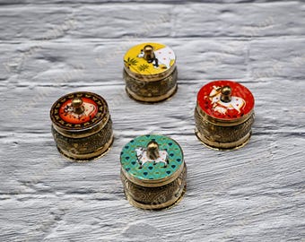 May include: Four small, round, decorative boxes with lids. The lids are painted with colorful designs, including a cow, and are topped with a small gold knob. The boxes have a gold-colored, ornate metal base. The boxes are on a light gray wooden surface.