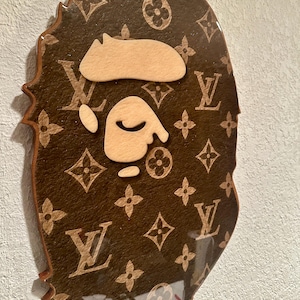 May include: A wall-mounted plaque shaped like a stylized ape head, featuring a brown surface with a repeating tan monogram pattern. The center of the plaque displays a light beige, smooth, cutout face and ear detail, all finished with a glossy coating.