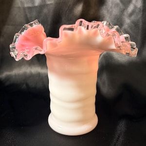 May include: A vintage glass vase with a ruffled rim. The vase features a gradient of colors, transitioning from white at the base to a soft pink at the top. The rim has a clear, textured edge. The vase has a cylindrical shape with a ridged texture.