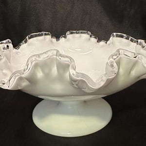 May include: A white milk glass pedestal bowl with a silver rim. The bowl has a ruffled edge and a round pedestal base.