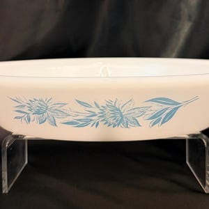 Könnte beinhalten: Eine ovale Auflaufform aus weißem Milchglas mit einem blauen Blumenmuster an der Außenseite. Die Form hat einen breiten Rand und steht auf einem durchsichtigen Acrylständer, der die glatte Oberfläche und das detaillierte blaue Blumenmotiv zeigt.
