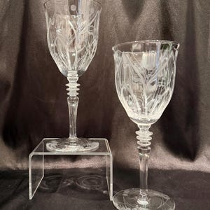 Könnte beinhalten: Zwei klare, geätzte Weingläser mit floralen und Blattmustern. Die Gläser haben einen detaillierten Stiel mit einem bauchigen Abschnitt und einem breiten, kreisförmigen Fuß. Ein Glas steht auf einem transparenten Acrylständer. Der Hintergrund ist dunkel.