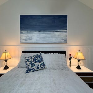 May include: Bedroom interior featuring a bed with a white fluffy comforter and pillows. A blue and white patterned pillow rests on the bed. Above the bed is a framed abstract painting in shades of blue and white. Two lamps with beige shades sit on nightstands.