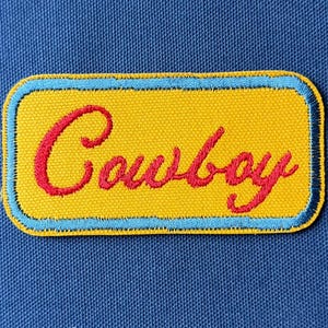 Puede incluir: Parche bordado amarillo con la palabra "Cowboy" en letras rojas. El parche tiene un borde azul claro y está sobre un fondo de tela azul. El parche es rectangular con esquinas redondeadas.