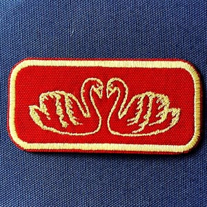 Puede incluir: Un parche rectangular rojo con un borde dorado y un diseño bordado en oro de dos cisnes enfrentados. El parche está sobre un fondo de tela azul.