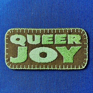 May include: A rectangular patch with the words "QUEER JOY" embroidered in light green on a brown background. The patch has a stitched border and is set against a blue fabric.