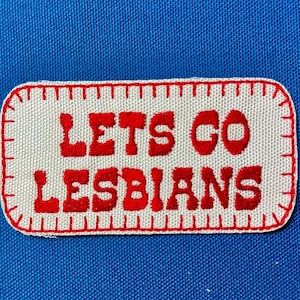 Könnte beinhalten: Ein rechteckiger Aufnäher mit den roten Worten "LETS GO LESBIANS", auf weißem Hintergrund aufgenäht. Der Aufnäher hat einen rot genähten Rand und befindet sich vor einem blauen Stoffhintergrund.