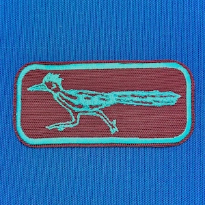 Peut inclure: Un patch en tissu marron avec un contour bleu sarcelle. Le patch présente une image brodée d'un coureur des routes, un oiseau du désert, avec une longue queue et un bec pointu.