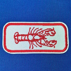 Puede incluir: Diseño de langosta bordada en rojo sobre un parche rectangular blanco con borde rojo.