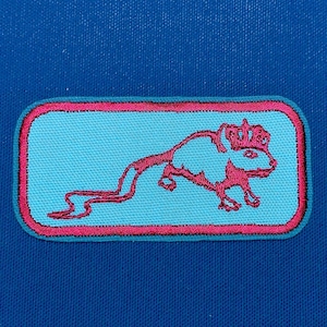 Puede incluir: Un parche bordado rosa con fondo azul. El parche presenta una rata de dibujos animados con una corona y una cola larga.