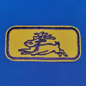 May include: A yellow rectangular patch with a purple embroidered outline. Inside the patch is a purple silhouette of a running rabbit with a bushy tail.