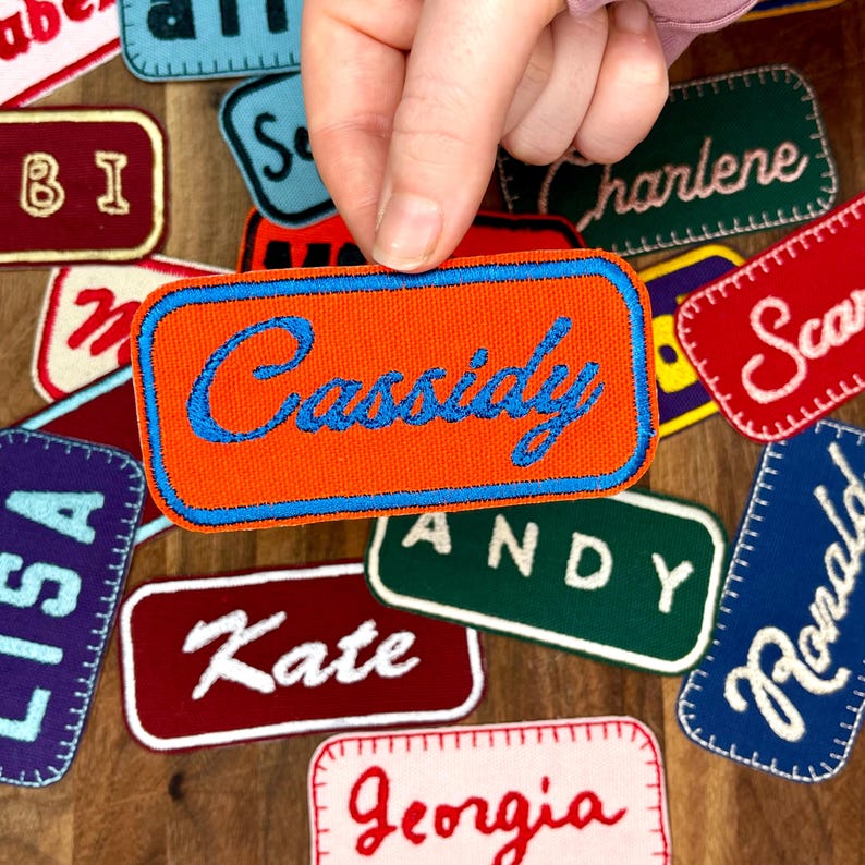 May include: Orange fabric patch with blue embroidered text that reads "Cassidy". The patch is surrounded by other fabric patches with names embroidered on them.