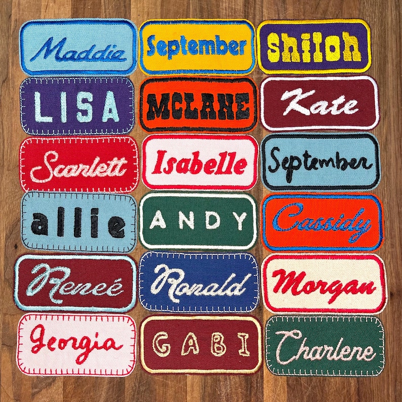 May include: A collection of 15 colorful fabric patches with names stitched on them. The patches are rectangular and have a white background with colorful stitching around the edges. The names on the patches include Maddie, September, Shiloh, Lisa, McLane, Kate, Scarlett, Isabelle, September, Allie, Andy, Cassidy, Renee, Ronald, Morgan, Georgia, Gabi, and Charlene.