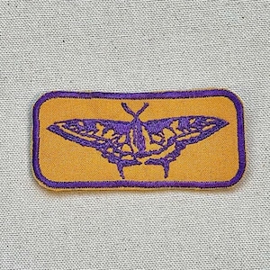 Op de afbeelding: Paarse geborduurde vlinder op een gele rechthoekige patch.