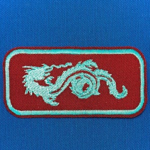 Könnte beinhalten: Ein gestickter Aufnäher in Rotbraun mit einem Design eines türkisfarbenen Drachen. Der Aufnäher hat einen türkisfarbenen Rand.