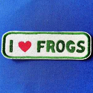 Pode incluir: Remendo bordado com o texto "I ♥ FROGS". O remendo apresenta um coração vermelho, texto verde e uma borda verde sobre um fundo branco. O remendo é retangular e tem cantos arredondados.