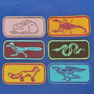 May include: Six colourful embroidered patches featuring various animals. The patches are rectangular and have a white background with a black outline. The animals include a rabbit, scorpion, snake, roadrunner, fox, and a bird.