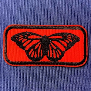 May include: A rectangular patch with a red background and a black butterfly design. The butterfly is centered within the patch, and the edges are outlined in black. The patch is set against a blue fabric background.