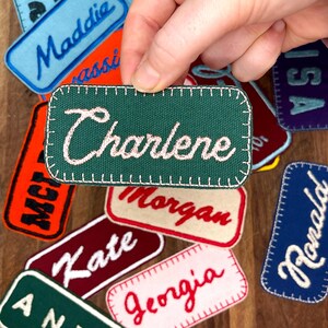 May include: A collection of personalized embroidered patches with various names, including Charlene, Maddie, Morgan, Kate, Georgia, and Ronald. The patches are made of green, blue, red, white, and black fabric.