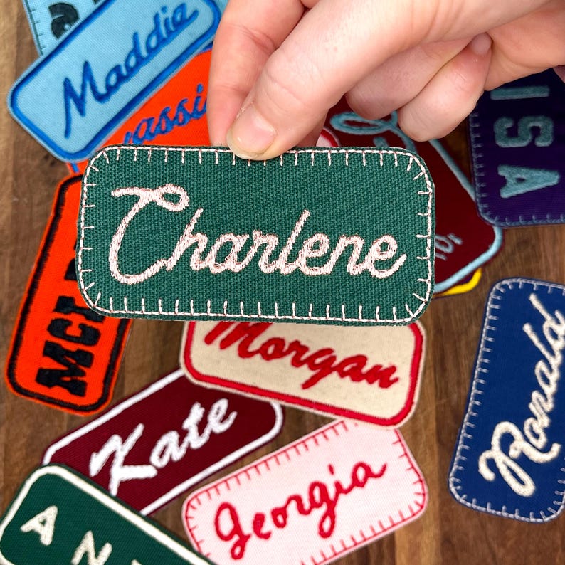 May include: A collection of personalized embroidered patches with various names, including Charlene, Maddie, Morgan, Kate, Georgia, and Ronald. The patches are made of green, blue, red, white, and black fabric.