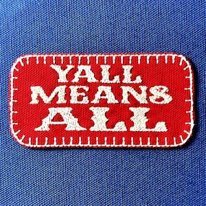 May include: A red patch with white embroidered text that reads "YALL MEANS ALL." The patch has a white stitched border and is set against a blue fabric background. The patch is rectangular with rounded corners.
