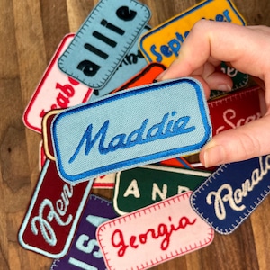 May include: A collection of colorful fabric patches with embroidered names, including "Allie", "Maddie", "Georgia", "Ren", "Ronald", and "September".