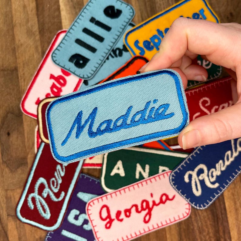 May include: A collection of colorful fabric patches with embroidered names, including "Allie", "Maddie", "Georgia", "Ren", "Ronald", and "September".