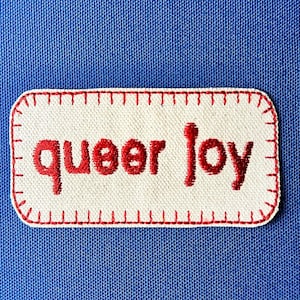 May include: A rectangular patch with the words "queer joy" embroidered in red on a white background. The patch has a red stitched border and is set against a blue fabric backdrop.
