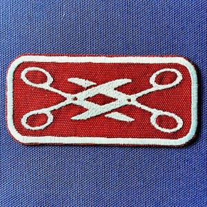 Pode incluir: Um patch retangular vermelho com uma borda branca e um design de tesoura branca. As tesouras são cruzadas no centro. O patch está em um fundo de tecido azul.