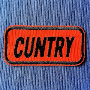 May include: An orange rectangular patch with the word "CUNTRY" in black, outlined in black. The patch is set against a blue fabric background.