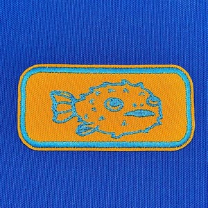 May include: A blue and orange embroidered patch featuring a cartoon pufferfish with a spiky exterior. The patch is rectangular and has a blue border.