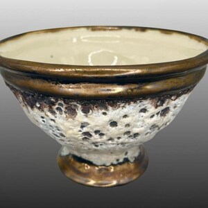 May include: A small white ceramic bowl with a textured surface and a gold rim. The bowl has a wide base and a rounded shape.