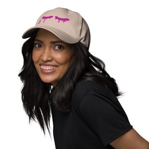 May include: A beige baseball cap with a curved peak. The cap has a magenta graphic of stylized eyelashes. The cap is made of a soft material and is suitable for everyday wear.